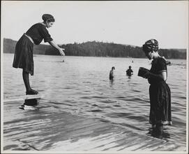 [Woman standing a lake, photographing another woman who is perched to dive]