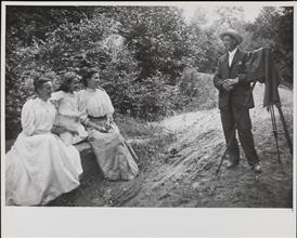 [Photographer with tripod set up to take a portrait of two women and a girl]