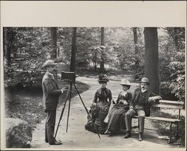 [Photographer with tripod set up to take a portrait of two women and a man]