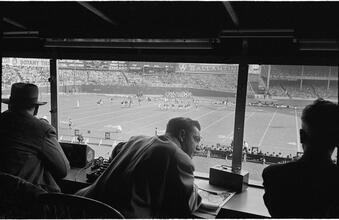 Football Writers [Reporters commentating on a football game in a stadium from the press box.]