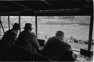 Football Writers [Reporters commentating on a football game in a stadium from the press box.]