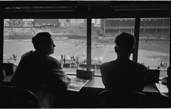Football Writers [Reporters commentating on a football game in a stadium from the press box.]