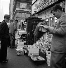 Newsstand | [People browsing through magazine racks at a busy sidewalk newsstand.]