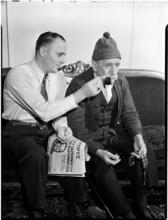 [Man lighting another man's pipe.]