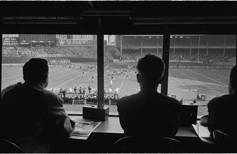 Football Writers [Reporters commentating on a football game in a stadium from the press box.]
