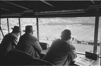 Football Writers [Reporters commentating on a football game in a stadium from the press box.]