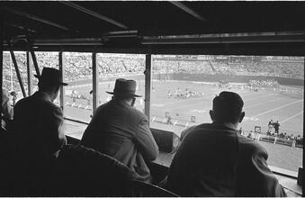 Football Writers [Reporters commentating on a football game in a stadium from the press box.]