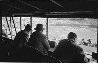 Football Writers [Reporters commentating on a football game in a stadium from the press box.]