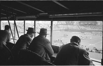 Football Writers [Reporters commentating on a football game in a stadium from the press box.]