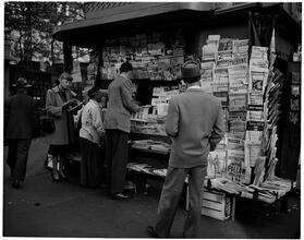 Newsstand [People browsing through magazine racks at a busy sidewalk newsstand.]
