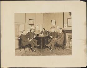 [Office interior, men gathered around a desk.]