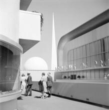[People walking by GM's Preview of Progress Auditorium at the New York World's Fair, with the Perisphere and Trylon in the background.]