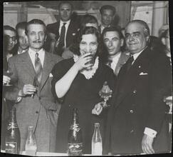 [Berta Gersten drinking a glass of champagne with a group of men.]