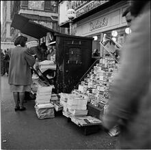 Newsstand | [People browsing through magazine racks at a busy sidewalk newsstand.]